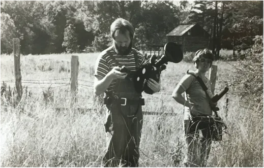 In a field, Peter Adair looks at light meter, holding a film camera, Lucy Phenix holds a recorder and mic.