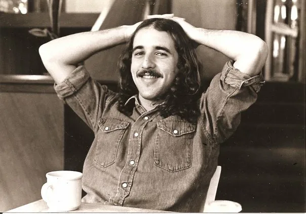 A long haired, moustached  and smiling young David Weismann relaxing at a cafe table.